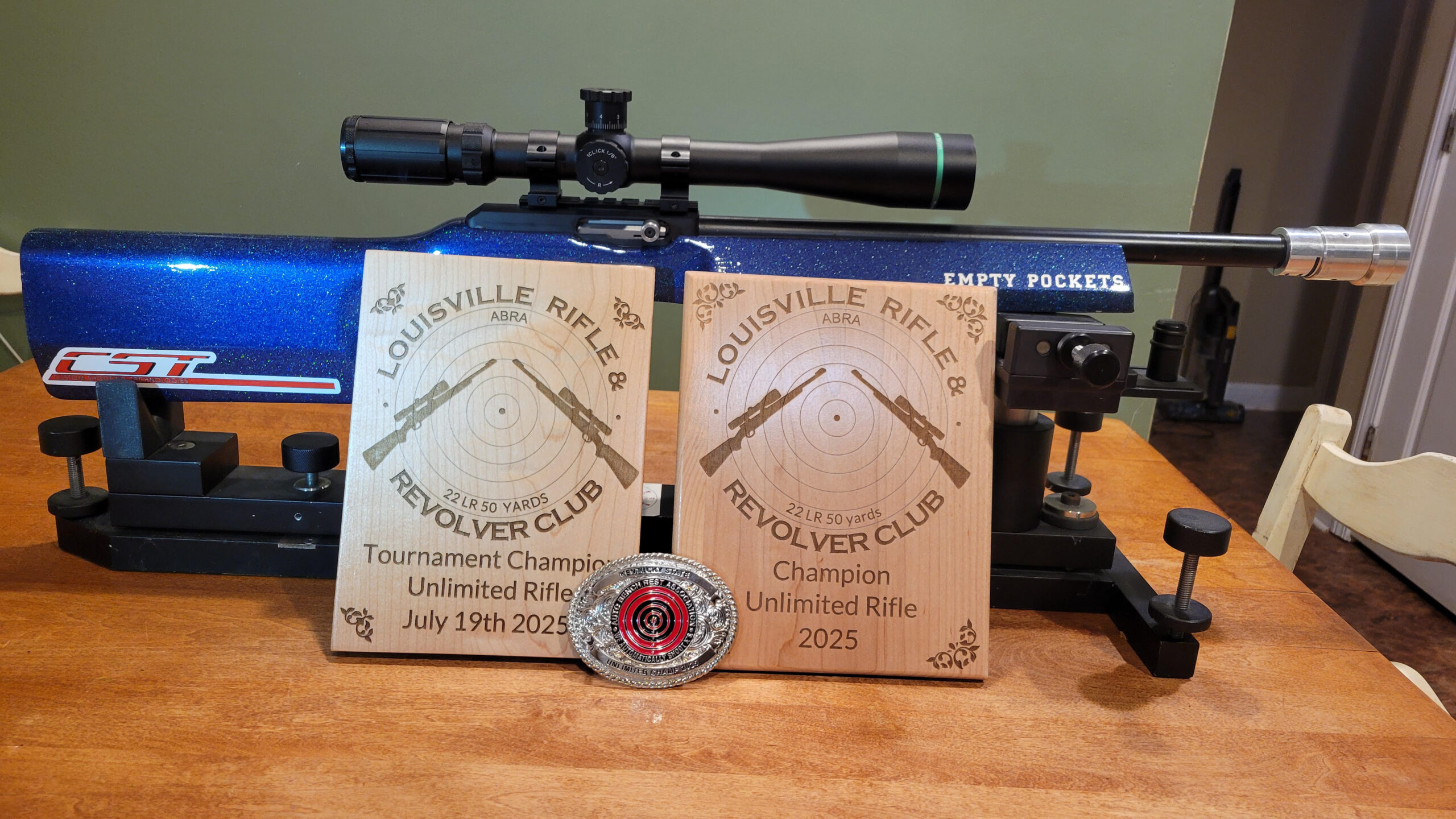 Glittery blue air rifle with a scope on a bench, flanked by two Louisville Rifle & Revolver Club target plaques and a silver medal on a wooden table.